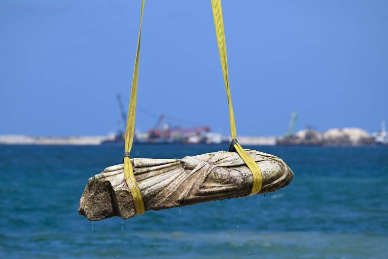 A imagem mostra uma estátua de pedra sendo içada do mar por meio de cintas amarelas. A estátua parece ter uma forma humana, mas está parcialmente submersa. Ao fundo, é possível ver o mar azul e algumas embarcações e estruturas portuárias. O céu está claro e ensolarado.