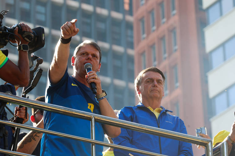 A imagem mostra um orador em um palanque, segurando um microfone e apontando com a mão. Ele está vestido com uma camiseta azul e amarela. Ao lado dele, há uma figura pública em um casaco azul, observando atentamente. O fundo apresenta edifícios altos e uma multidão visível.