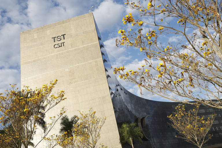 A imagem mostra o prédio do Tribunal Superior do Trabalho (TST) no Brasil, com uma estrutura moderna e angular. O edifício é de cor clara e possui a sigla 'TST' e 'CSJT' visíveis na fachada. Em primeiro plano, há árvores com flores amarelas, e o céu está parcialmente nublado.