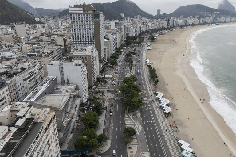 A imagem mostra uma vista aérea da praia de Copacabana, no Rio de Janeiro. À esquerda, há uma série de edifícios, enquanto à direita se vê a praia com areia clara e algumas pessoas. A orla é acompanhada por uma avenida com árvores e calçadas. Ao fundo, montanhas e um céu nublado completam a cena.