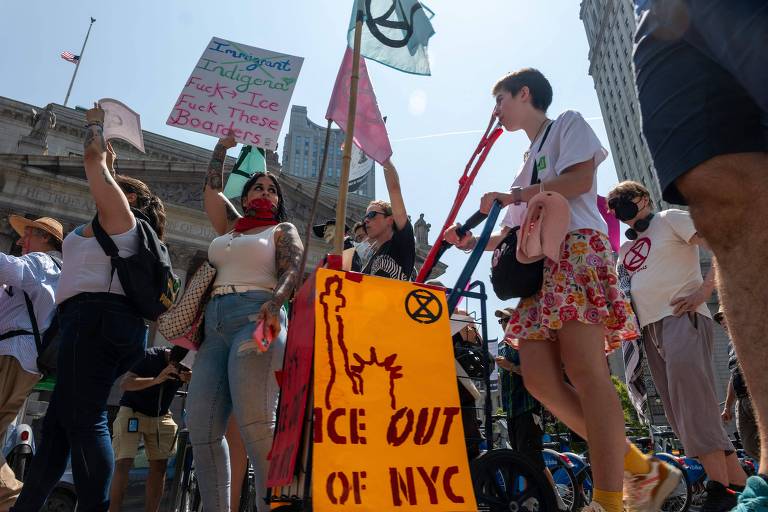 A imagem mostra um grupo de manifestantes em um protesto ao ar livre. Algumas pessoas seguram cartazes, incluindo um que diz 'ICE OUT OF NYC'. Os manifestantes estão vestidos de maneira variada, com algumas pessoas usando máscaras. O cenário é urbano, com edifícios ao fundo e um céu claro.