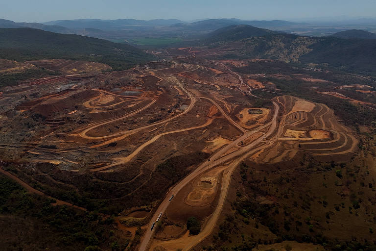 A imagem mostra uma vasta área de mineração, com terrenos escavados e estradas de acesso. O solo apresenta diferentes tonalidades de marrom e laranja, indicando a remoção de camadas de terra. Ao fundo, há montanhas e uma paisagem verdejante, contrastando com a área desmatada e alterada pela atividade mineradora.