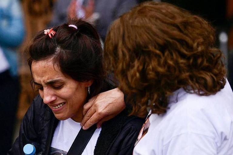De afbeelding toont twee vrouwen in een emotioneel moment. De vrouw links, met donker, opgestoken haar, huilt en lijkt diep bedroefd. De vrouw rechts, met los, bruin haar, troost de vrouw links en legt haar hand op haar schouder. De achtergrond is wazig, maar er zijn andere mensen zichtbaar, wat duidt op een emotioneel ondersteunende omgeving.