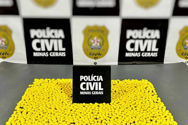 De afbeelding toont een groot aantal gele pillen, gerangschikt op een oppervlak. In het midden staat een zwart bord met de tekst 'POLÍCIA CIVIL MINAS GERAIS'. Op de achtergrond zijn meer borden van de civiele politie zichtbaar, die de inbeslagname markeren.