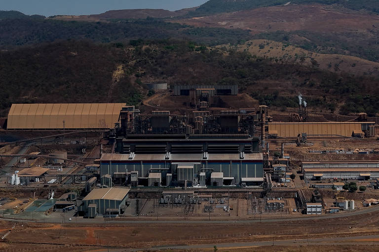 A imagem mostra uma vista aérea de uma grande instalação industrial, com várias estruturas metálicas e edifícios. No centro, há uma construção principal com várias chaminés e janelas. Ao fundo, é possível ver uma área montanhosa e vegetação escassa. A área ao redor da indústria parece ser predominantemente árida, com algumas estruturas menores e equipamentos visíveis.