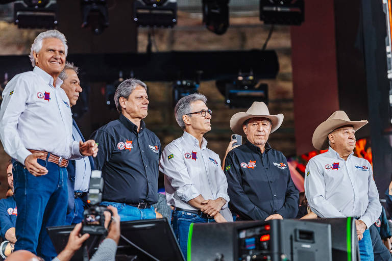 A imagem mostra um grupo de seis homens em um palco durante um evento político. Eles estão vestidos com camisas de diferentes cores, algumas com logotipos visíveis. O homem à direita usa um chapéu de cowboy. Ao fundo, há uma iluminação que sugere um ambiente festivo ou de celebração. Na frente, há uma câmera focada nos homens, indicando que o evento está sendo filmado ou transmitido.