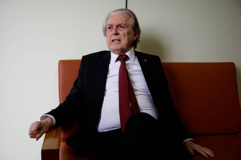 Um homem idoso está sentado em um sofá de couro marrom. Ele usa um terno escuro com uma gravata vermelha e tem cabelo grisalho. O fundo é uma parede clara, e a iluminação é suave. O homem parece estar em uma conversa ou reflexão, com uma expressão séria.
