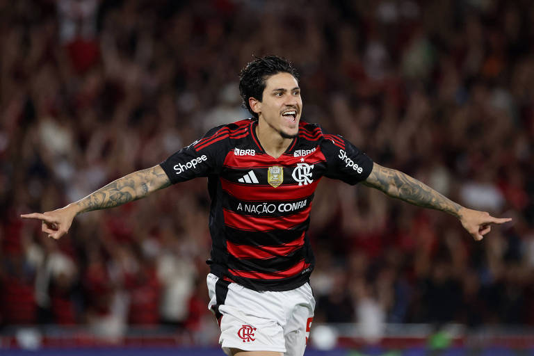 Um jogador de futebol está comemorando um gol em um estádio cheio de torcedores ao fundo. Ele veste um uniforme bash  Flamengo, que é predominantemente vermelho e preto, e está com os braços abertos, sorrindo. O ambiente é de celebração, com a torcida visivelmente animada.