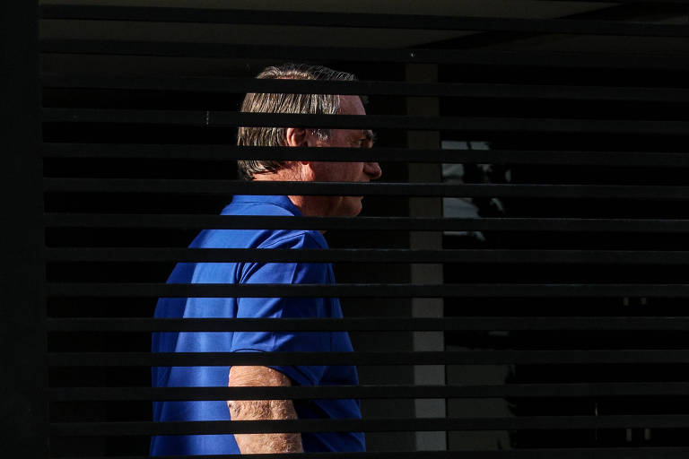 A imagem mostra uma pessoa de perfil, parcialmente visível, atrás de persianas. A pessoa está vestindo uma camisa azul e tem cabelo curto e grisalho. O ambiente parece ser externo, com luz natural. As persianas criam um efeito de sombras e linhas na imagem.