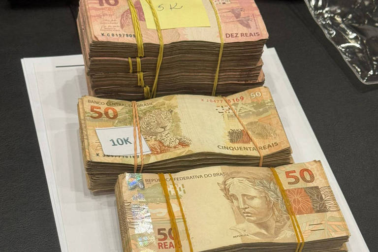 A imagem mostra três pilhas de notas de dinheiro empilhadas sobre uma superfície branca. As notas são de diferentes denominações, com a pilha maior contendo notas de valor mais alto. Ao fundo, é possível ver telefones e um plástico transparente.