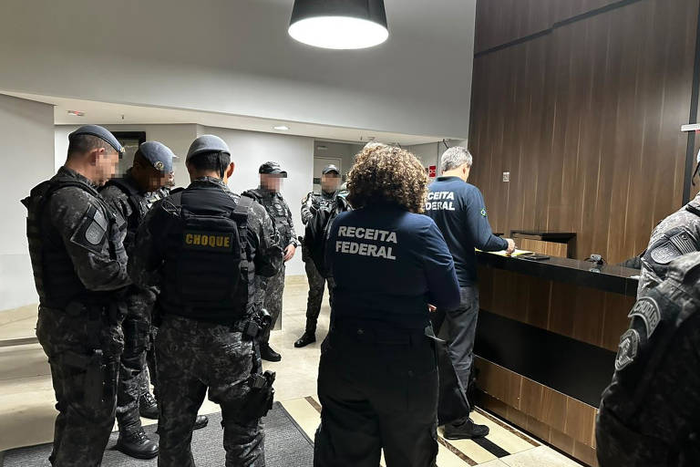 A imagem mostra um grupo de policiais em um ambiente interno, possivelmente um hotel. Os policiais estão vestidos com uniformes táticos e alguns usam bonés. Eles estão em pé, em volta de uma mesa de recepção, onde um homem está interagindo com eles. O ambiente é bem iluminado, com paredes de madeira e uma iluminação pendente visível no teto.