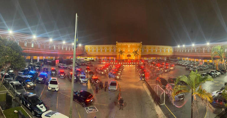 A imagem mostra um grande estacionamento iluminado à noite, com várias luzes brilhantes. Há muitos carros estacionados, incluindo veículos de emergência com luzes piscando. Ao fundo, pode-se ver um edifício grande e iluminado, que parece ser uma instituição pública ou um centro de eventos. O ambiente é movimentado, com várias pessoas visíveis no estacionamento.