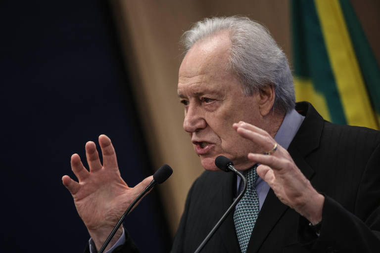 Um homem de cabelos grisalhos e com um terno escuro está falando em um microfone. Ele gesticula com as mãos, expressando-se de forma enfática. Ao fundo, há uma bandeira do Brasil visível, com as cores verde e amarelo.