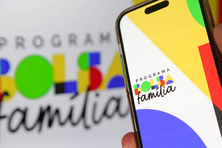 A imagem mostra um smartphone exibindo a tela do aplicativo do Programa Bolsa Família. O fundo da tela é colorido, com formas geométricas em amarelo, verde, azul e vermelho. O logotipo do programa está em destaque, com as palavras 'PROGRAMA BOLSA FAMÍLIA' em uma fonte estilizada. Ao fundo, é possível ver uma versão ampliada do logotipo em uma superfície plana.