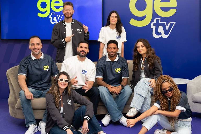 Um grupo de sete pessoas posando para a foto em um estúdio. Eles estão vestindo camisetas e jaquetas com a marca 'ge'. O fundo é azul com o logotipo 'ge tv' em destaque. As pessoas estão em diferentes poses, algumas sorrindo e outras com expressões descontraídas. O ambiente é moderno e bem iluminado.