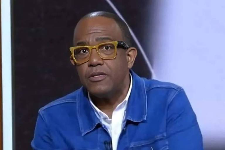 Um homem está sentado, olhando para a câmera. Ele usa uma jaqueta jeans azul e uma camisa branca por baixo. O homem tem cabelo curto e usa óculos de armação amarela. O fundo da imagem é desfocado, mas parece ser um ambiente de estúdio.