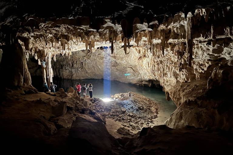 A imagem mostra o interior de uma caverna com formações de estalactites e estalagmites. Há um grupo de pessoas em pé, observando a caverna, e um feixe de luz que ilumina uma área com água. As paredes da caverna são rochosas e irregulares, e o ambiente é escuro, exceto pela luz que destaca a beleza natural do local.
