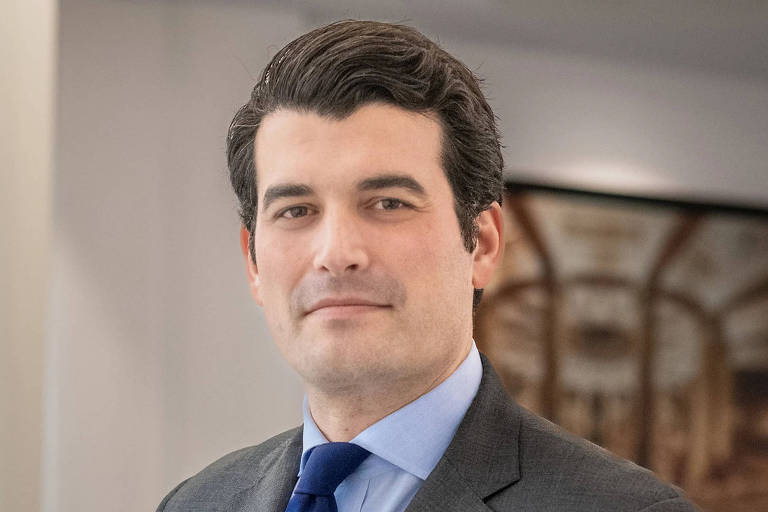 Um homem está posando para a foto, vestindo um terno cinza e uma camisa azul com uma gravata azul. Ele tem cabelo escuro e liso, e está em um ambiente interno com uma decoração neutra ao fundo. O homem tem uma expressão séria e confiante.