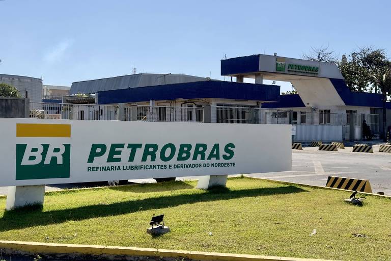 A imagem mostra a entrada de uma refinaria da Petrobras, com um grande letreiro em primeiro plano. O letreiro exibe o logotipo da empresa, que inclui as letras 'BR' e a palavra 'PETROBRAS', além da descrição 'REFINARIA LUBRIFICANTES E DERIVADOS DO NORDESTE'. Ao fundo, é possível ver a estrutura da refinaria, com um céu claro e algumas árvores ao redor.
