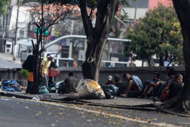 A imagem mostra um grupo de pessoas em situação de rua em um ambiente urbano. Algumas pessoas estão sentadas no chão, enquanto outras estão de pé. Há sacos de lixo e cobertores espalhados pelo chão. Árvores estão presentes ao redor, e ao fundo, é possível ver edifícios e uma rua. A iluminação sugere que é um dia claro.