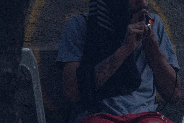 A imagem mostra um homem sentado em uma cadeira plástica, fumando um cigarro. Ele está usando uma camiseta azul e calças vermelhas. O homem tem um lenço ou toalha cobrindo parte da cabeça e está em um ambiente urbano, com uma parede ao fundo que possui marcas ou grafites. A iluminação é baixa, sugerindo que a foto foi tirada em um momento de pouca luz.