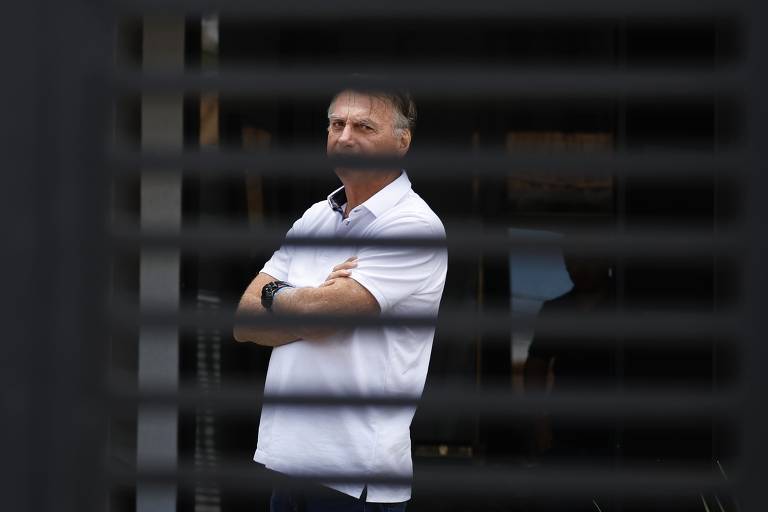 Um homem está em pé, com os braços cruzados, olhando para frente. Ele usa uma camisa branca e está posicionado atrás de uma grade ou persiana, que cria um efeito de linhas horizontais na imagem. O fundo é desfocado, mas parece haver uma área externa iluminada.
