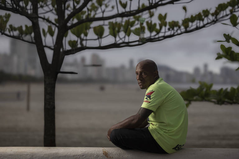 Um homem de pele escura está sentado em um muro à beira da praia, olhando para a câmera. Ele usa uma camiseta verde com detalhes em preto e vermelho. Ao fundo, há uma paisagem de praia com areia e um céu nublado, além de uma cidade visível à distância. Uma árvore com folhas verdes está próxima ao homem, criando uma sombra parcial sobre ele.