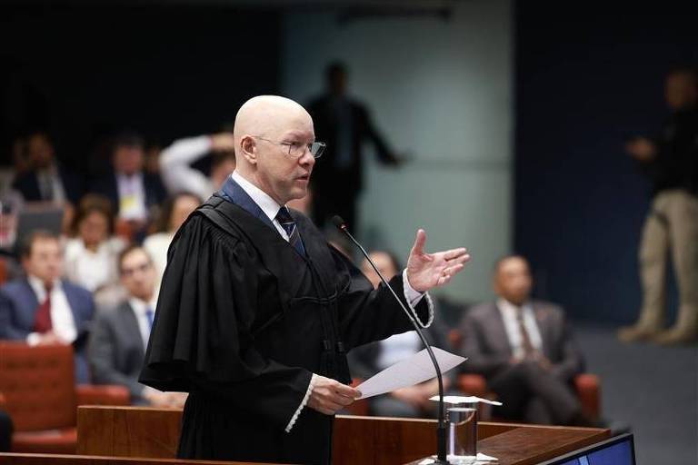 Um juiz careca, vestido com uma toga preta e gravata, está em pé em um tribunal, segurando um papel e gesticulando com a mão. Ao fundo, há várias pessoas sentadas, algumas em trajes formais, observando atentamente. O ambiente é bem iluminado, com um painel azul ao fundo e uma mesa de tribunal à frente do juiz.