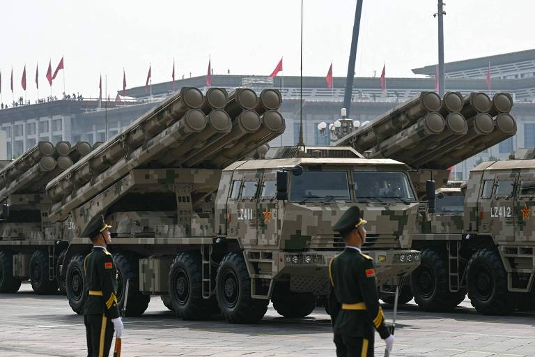 На снимке изображены военные ракетные пусковые установки на параде. Машины замаскированы в зелёно-коричневых тонах, сзади видны несколько пусковых труб. Перед машинами стоят по стойке смирно солдаты в парадной форме, головных уборах и с знаками различия. На заднем плане видно здание с красными флагами и толпа людей.