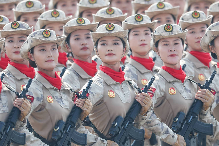 На снимке изображена группа женщин на военном параде, все в камуфляжной форме и головных уборах. Они выстроились в шеренгу, с серьёзными лицами и винтовками в руках. У каждой на шее красный шарф, а на форме видны эмблемы.