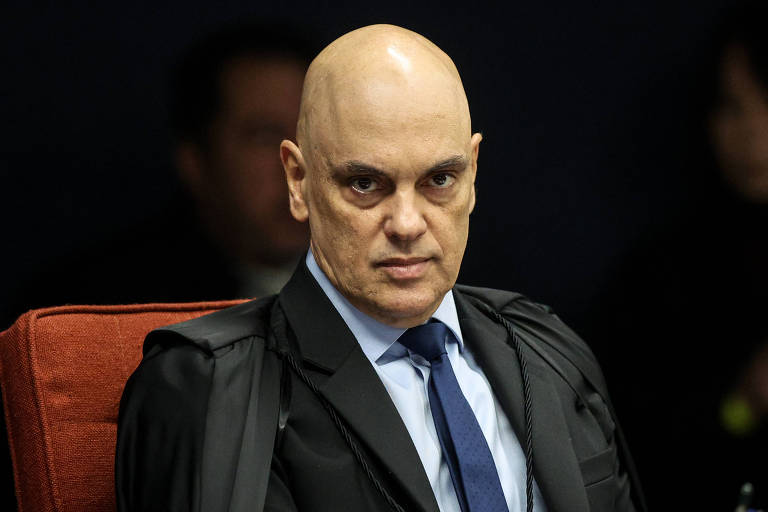 Um homem careca, com expressão séria, está sentado em uma cadeira vermelha. Ele usa um terno escuro com uma gravata azul e uma toga preta. Ao fundo, é possível ver algumas pessoas desfocadas, mas não é possível identificar detalhes.