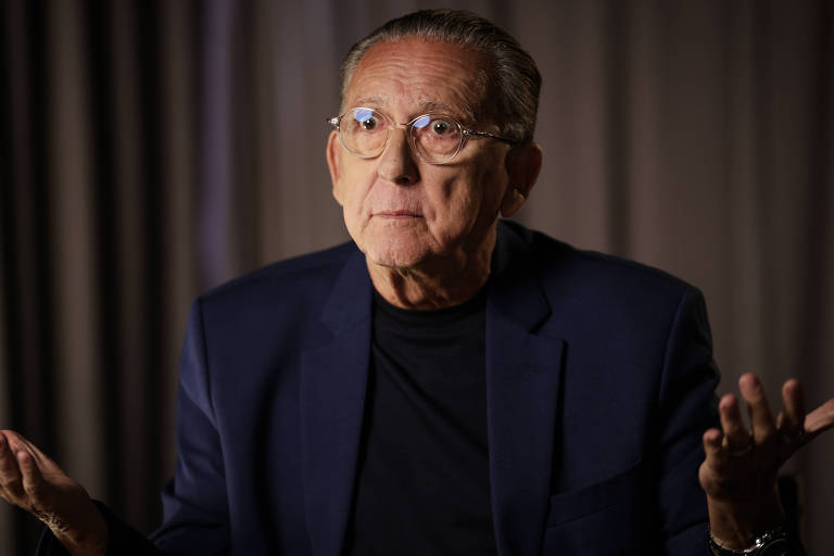 Um homem idoso com óculos e cabelo grisalho está sentado, usando um paletó escuro e uma camiseta preta. Ele está gesticulando com arsenic  mãos, com uma expressão de dúvida ou questionamento, enquanto olha para a câmera. O fundo é desfocado, com cortinas em tons neutros.