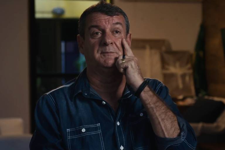 Um homem está sentado em um ambiente interno, com uma expressão pensativa. Ele usa uma camisa jeans de manga longa e está tocando o rosto com a mão. Ao fundo, há uma sala com móveis e uma televisão. A iluminação é suave, criando um ambiente acolhedor.