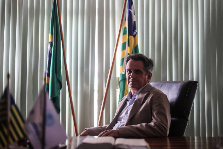 Um homem sentado em uma cadeira de escritório, com uma expressão séria. Ele está vestido com um paletó claro e uma camisa escura. Ao fundo, há cortinas verticais e várias bandeiras, incluindo bandeiras do Brasil e de estados. A mesa à sua frente tem um livro aberto.
