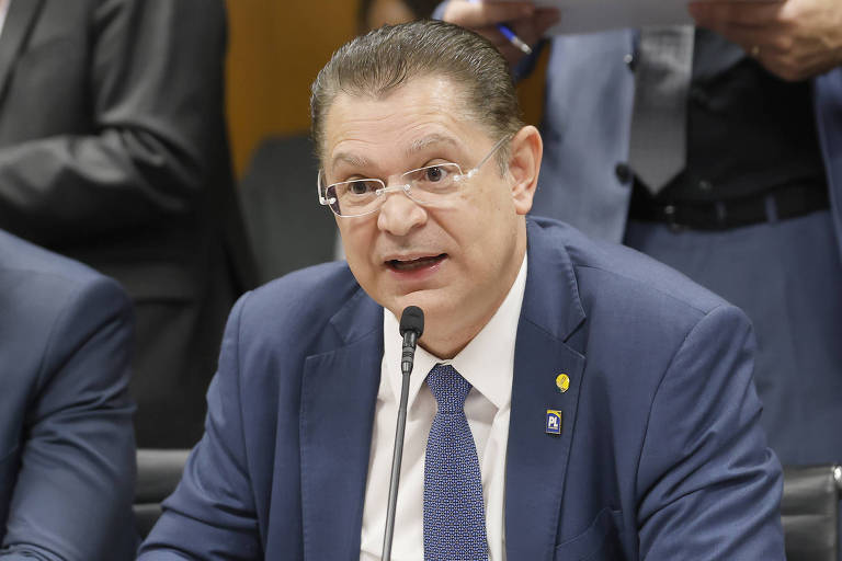 A bespectacled man, dressed in a dark suit and tie, speaks into a microphone during a public hearing. He appears to be explaining something important, with a serious expression. In the background, other people are seated, and one man is holding papers.