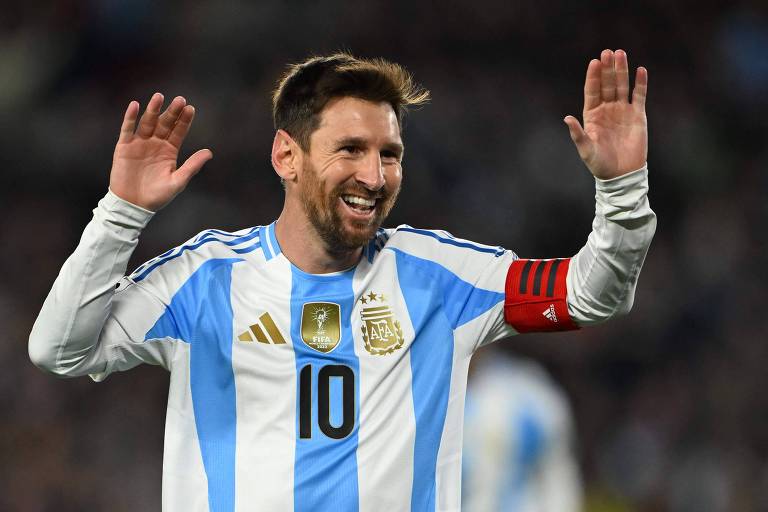 Um jogador de futebol está sorrindo e levantando arsenic  mãos em um gesto de comemoração. Ele veste uma camisa de futebol com listras verticais em azul e branco, com o número 10 nas costas. O jogador também usa uma braçadeira de capitão nary  braço esquerdo. O fundo mostra uma multidão em um estádio.