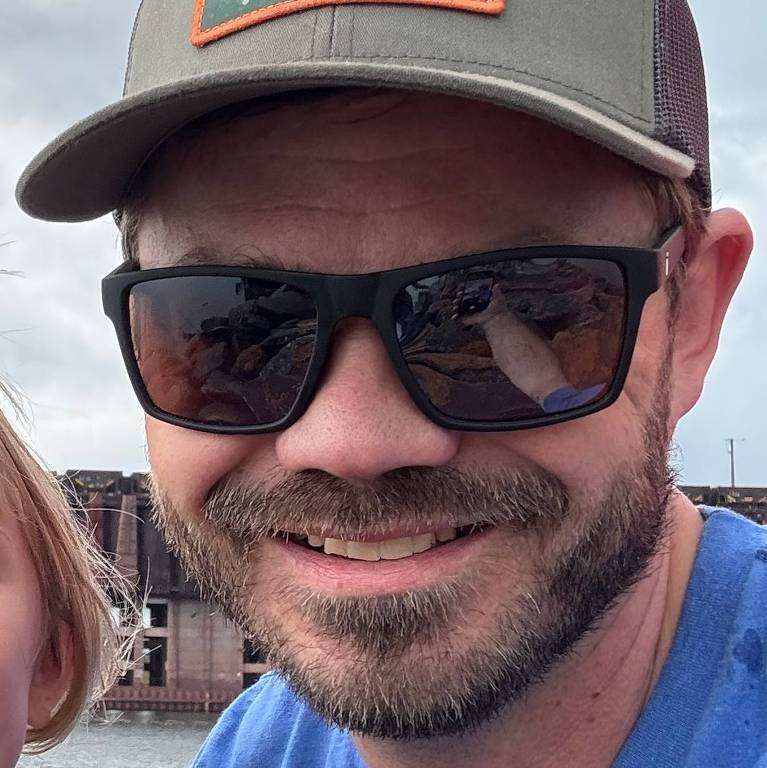 Um homem sorridente usando óculos escuros e um boné cinza com um emblema. Ele está vestindo uma camiseta azul e está em um ambiente ao ar livre, possivelmente perto de água. O fundo é desfocado, mas parece haver estruturas de madeira ao longe.