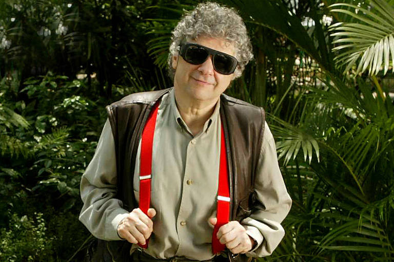 Um homem está encostado em uma grade verde em um parque. Ele usa óculos escuros, suspensórios vermelhos, uma camisa cinza e calças escuras. O fundo é composto por vegetação verde e árvores, criando um ambiente natural e tranquilo.