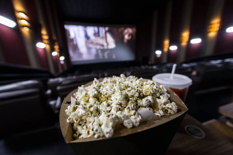 A imagem mostra uma caixa de pipoca cheia em primeiro plano, com um copo de bebida ao lado. Ao fundo, é possível ver uma tela de cinema com uma cena de filme sendo exibida. O ambiente é escuro, com poltronas de cinema visíveis e iluminação suave nas paredes.