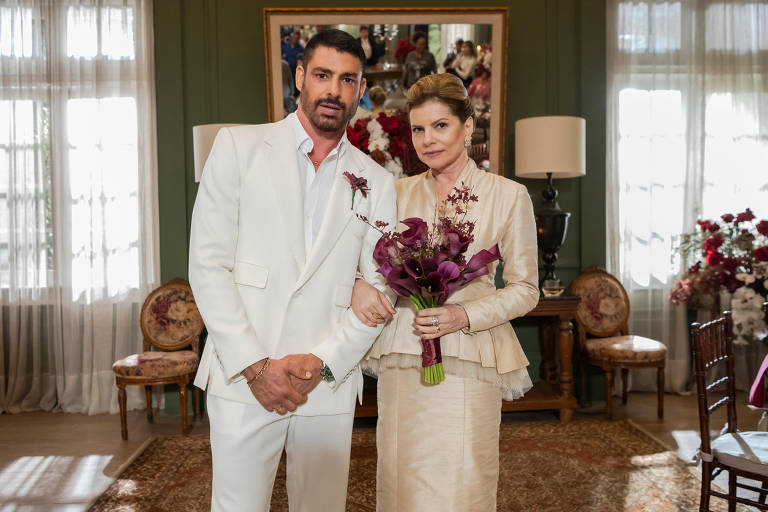 Um homem e uma mulher posam juntos em um ambiente elegante. O homem está vestido com um terno branco e tem cabelo escuro e barba. A mulher usa um vestido claro e segura um buquê de flores roxas. Ao fundo, há uma parede decorada com quadros e uma iluminação suave, além de móveis clássicos como cadeiras e mesas.