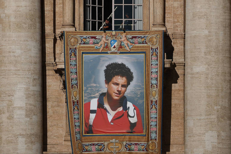 A imagem mostra um grande banner pendurado em uma estrutura arquitetônica, com um retrato de um jovem. O jovem tem cabelo encaracolado e está usando uma camiseta vermelha. O fundo do retrato apresenta um céu claro. O banner é decorado com um contorno ornamentado e está fixado em uma parede de pedra clara.