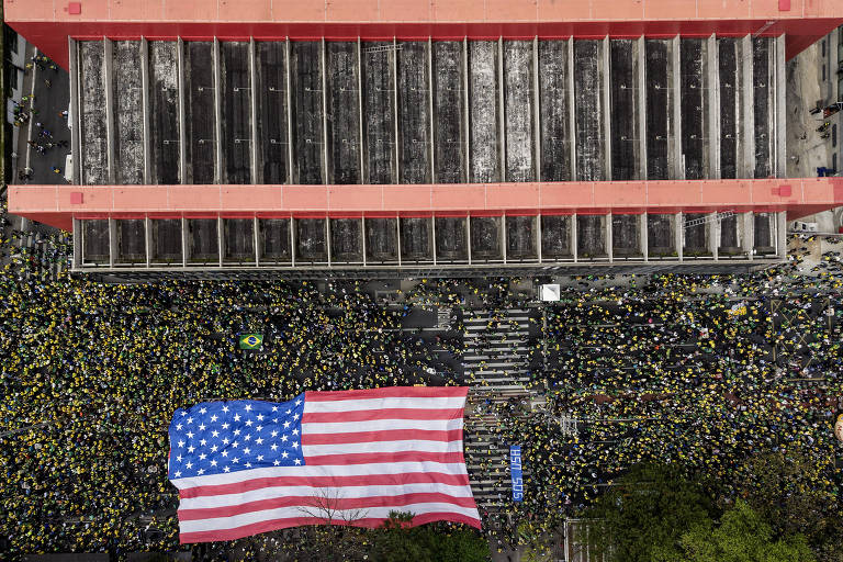 A imagem mostra uma grande multidão reunida em uma manifestação, vista de cima. A maioria das pessoas está vestindo roupas amarelas, e no centro da cena, há uma grande bandeira dos Estados Unidos estendida no chão. Ao fundo, pode-se ver um edifício com um telhado vermelho e uma estrutura de vidro. A bandeira do Brasil também é visível entre a multidão.