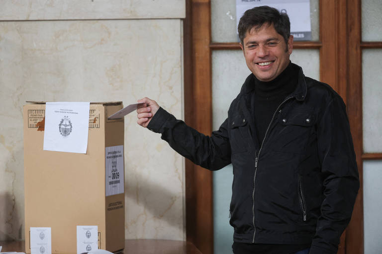 Um homem sorridente está colocando um papel em uma urna eleitoral. Ele está vestindo uma jaqueta preta e está em um ambiente interno, com paredes claras e janelas ao fundo. Ao lado da urna, há alguns papéis ou cédulas.