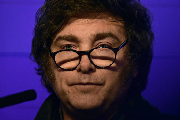 Um homem com cabelo castanho e ondulado, usando óculos redondos. Ele tem uma expressão séria e está em um ambiente com iluminação suave, que cria um fundo azul. O homem está vestido com uma jaqueta escura e parece estar em um evento ou apresentação.