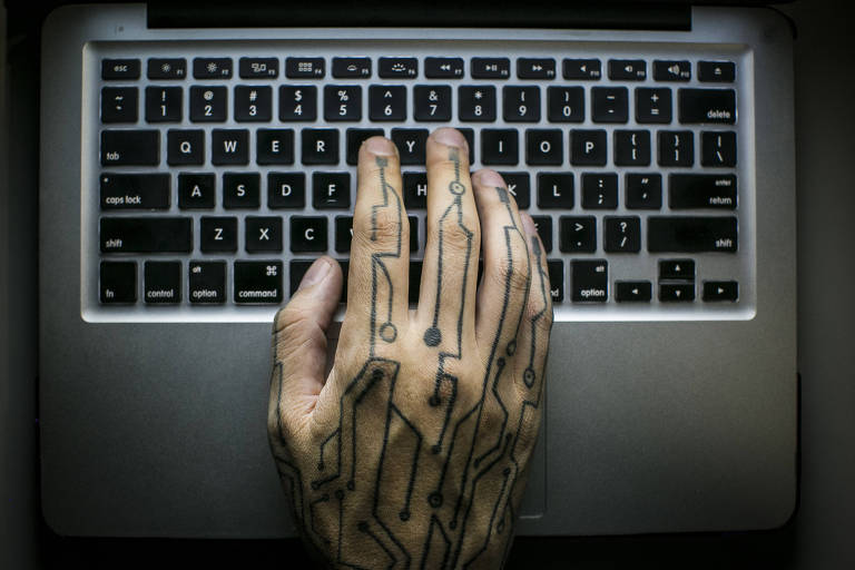 A imagem mostra uma mão com uma tatuagem de circuito eletrônico, posicionada sobre o teclado de um laptop. O teclado é de cor preta com letras brancas, e a mão está em uma posição que sugere que a pessoa está digitando. A iluminação é suave, destacando os detalhes da tatuagem e do teclado.
