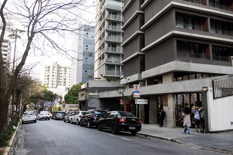 A imagem mostra uma rua urbana com edifícios modernos ao fundo. À esquerda, há uma fila de carros estacionados e, à direita, um prédio de vários andares com uma entrada visível. Algumas pessoas estão na calçada em frente ao prédio. Árvores sem folhas estão presentes ao longo da rua, e o céu está nublado.