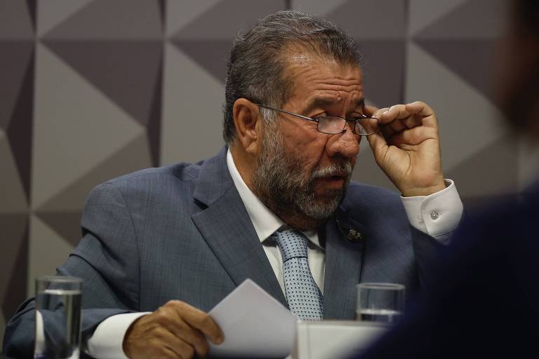 Um homem de barba e cabelo grisalho, usando um terno azul e uma gravata clara, está sentado à mesa em uma reunião. Ele está ajustando seus óculos e parece estar concentrado. Na mesa, há copos de água e papéis. O fundo apresenta um padrão geométrico em tons de cinza.