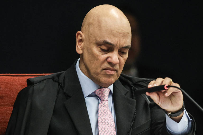 Um juiz com cabeça calva e expressão séria está sentado em uma cadeira, segurando um microfone. Ele usa um terno escuro e uma gravata rosa, com uma camisa clara. O fundo é escuro e há um detalhe em vermelho na cadeira.