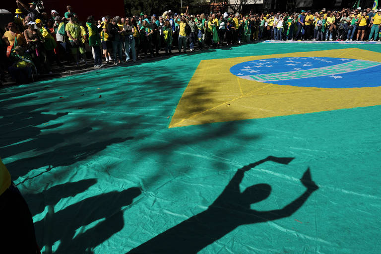 A imagem mostra uma grande bandeira do Brasil estendida no chão, com pessoas ao redor vestindo roupas nas cores verde e amarelo. A sombra de uma mão é projetada sobre a bandeira, formando um gesto de 'ok'. O ambiente parece ser de uma manifestação ou evento público.