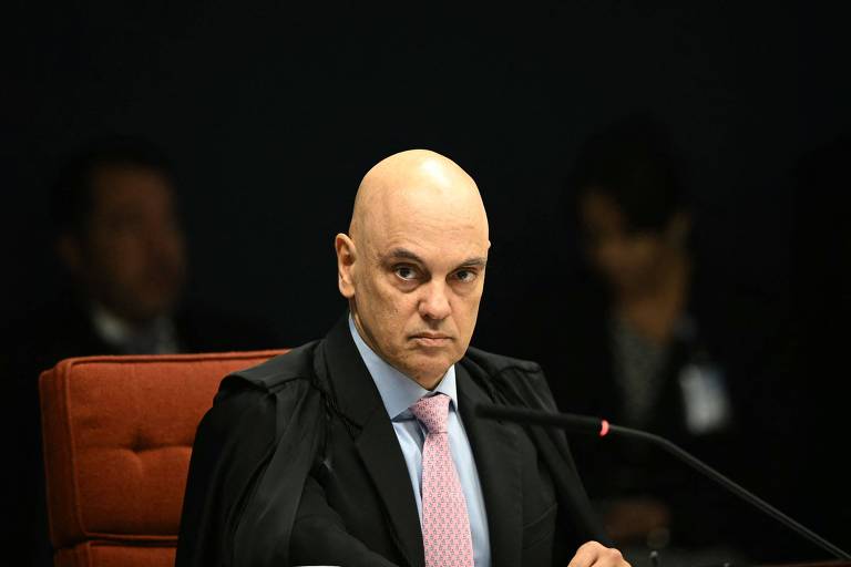 Um homem careca, vestido com uma toga preta e uma camisa clara, está sentado em uma mesa. Ele tem uma expressão séria e está olhando para frente. Ao fundo, há outras pessoas, mas não estão claramente visíveis. O ambiente parece ser uma sala de tribunal, com uma iluminação suave.
